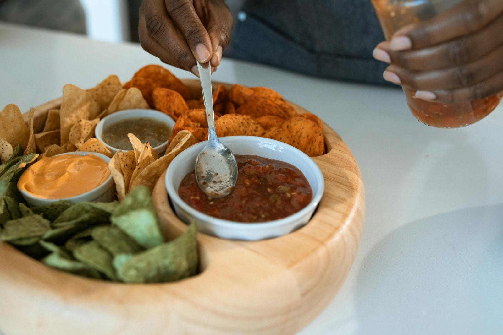 A hand serving salsa from a platter with assorted chips and dips on a kitchen counter.