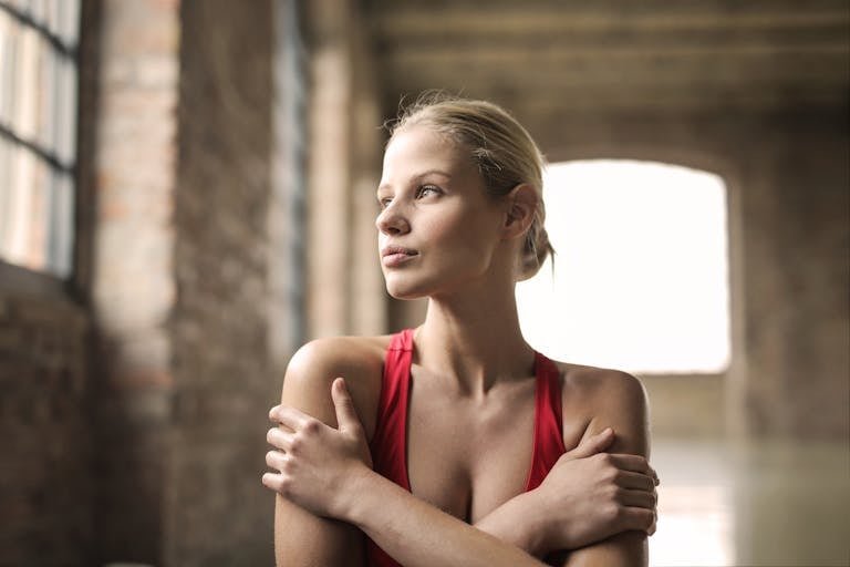 Serious millennial determined woman in sportswear looking away and thinking while standing alone and embracing self against blurred interior of loft studio